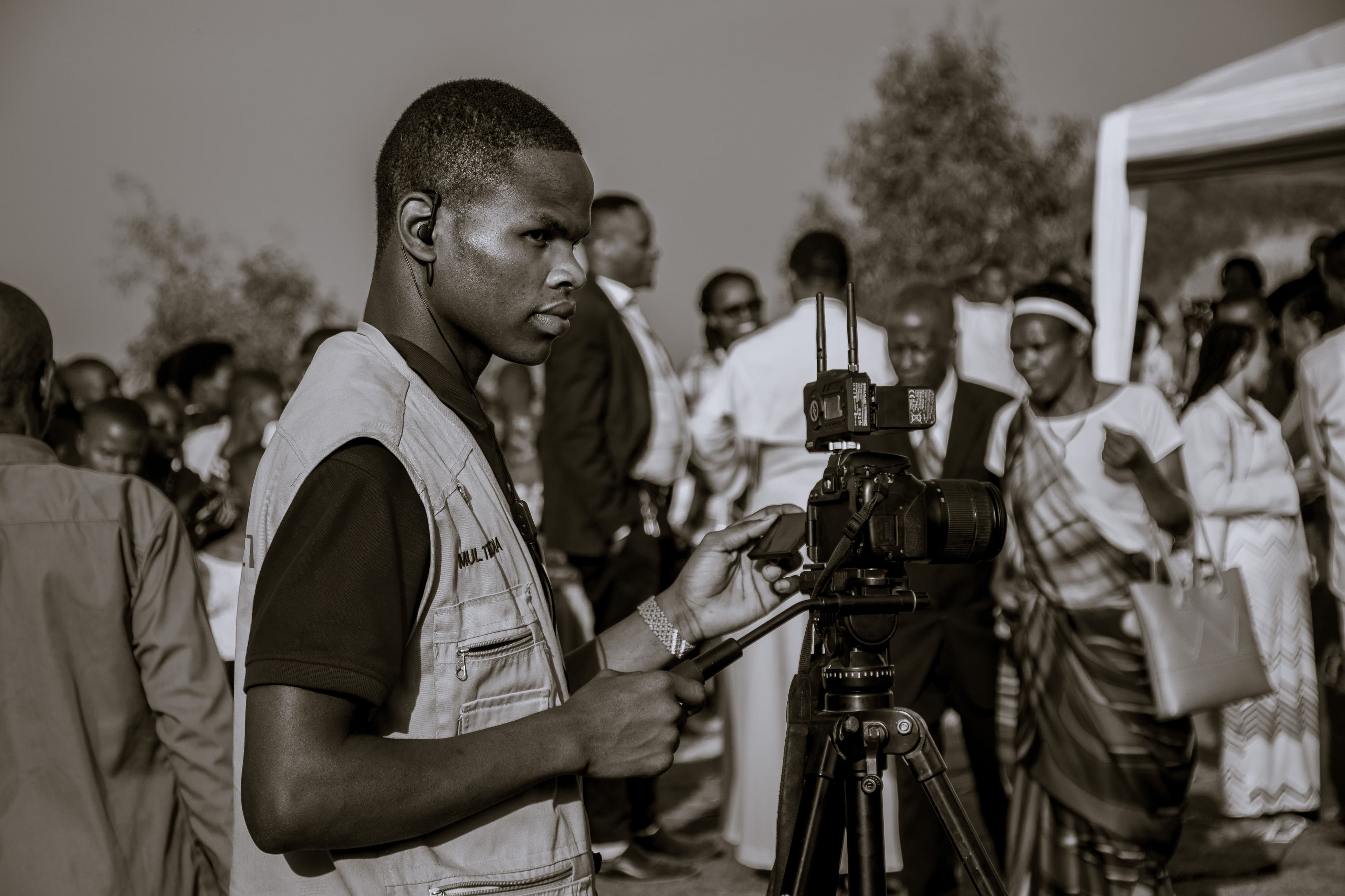Black-and-white photograph of a video operator in a reporter’s vest, an earpiece in his ear, framing a shot on a camera mounted on a tripod at an outdoor ceremony, a crowd of guests and a marquee behind him. — Photo: shutter Rwanda, "Photographer Capturing Event in Ruhango, Rwanda", via Pexels, Pexels License (https://www.pexels.com/license/)