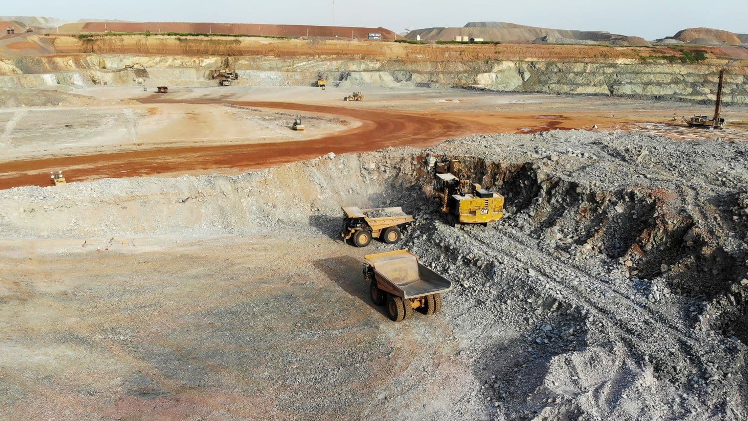 Aerial view of an open-pit mine: a yellow hydraulic excavator loading a haul truck at the foot of a working face, a second empty truck waiting on the pit floor, red laterite haul roads and benches behind.