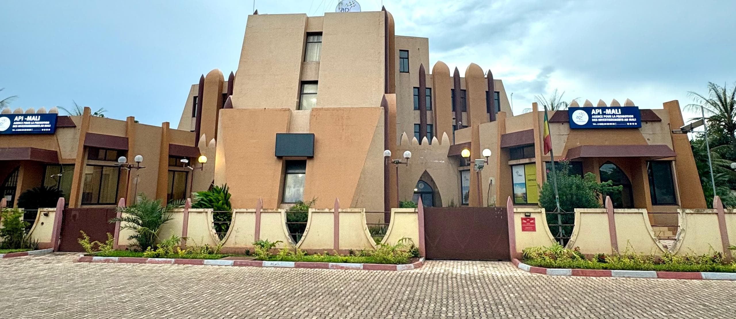 Front of the API-Mali headquarters in Bamako: a beige Sudano-Sahelian building with maroon pilasters, two “API-MALI” signboards, a Malian flag and a closed gate. — API-Mali
