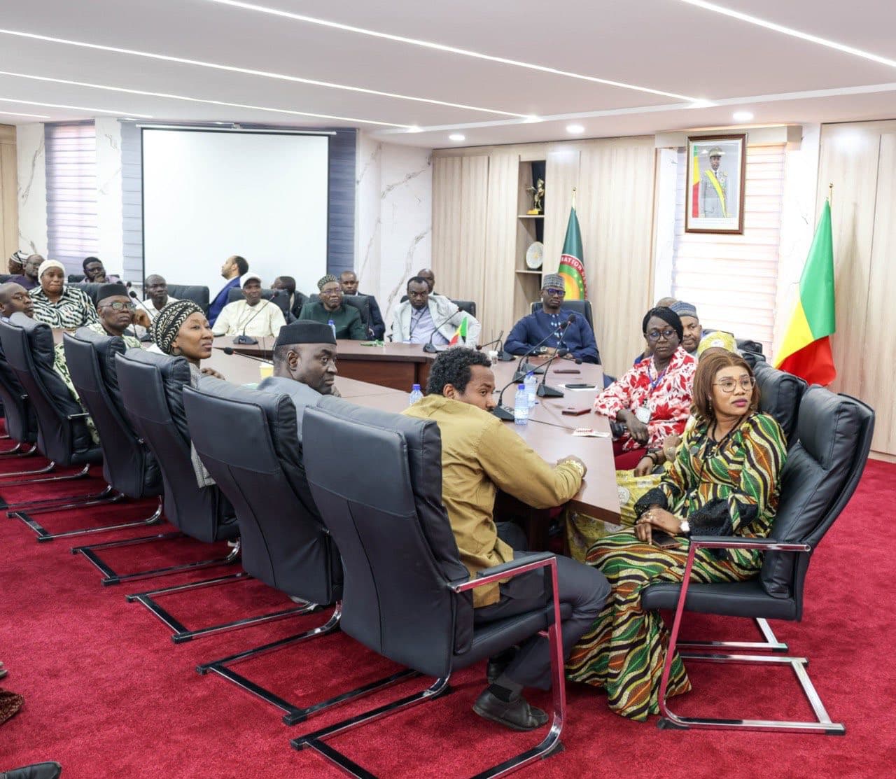 Large marble-clad official meeting room with two long rows of black chairs at microphone-fitted desks, and around twenty participants in boubous and suits. — API-Mali