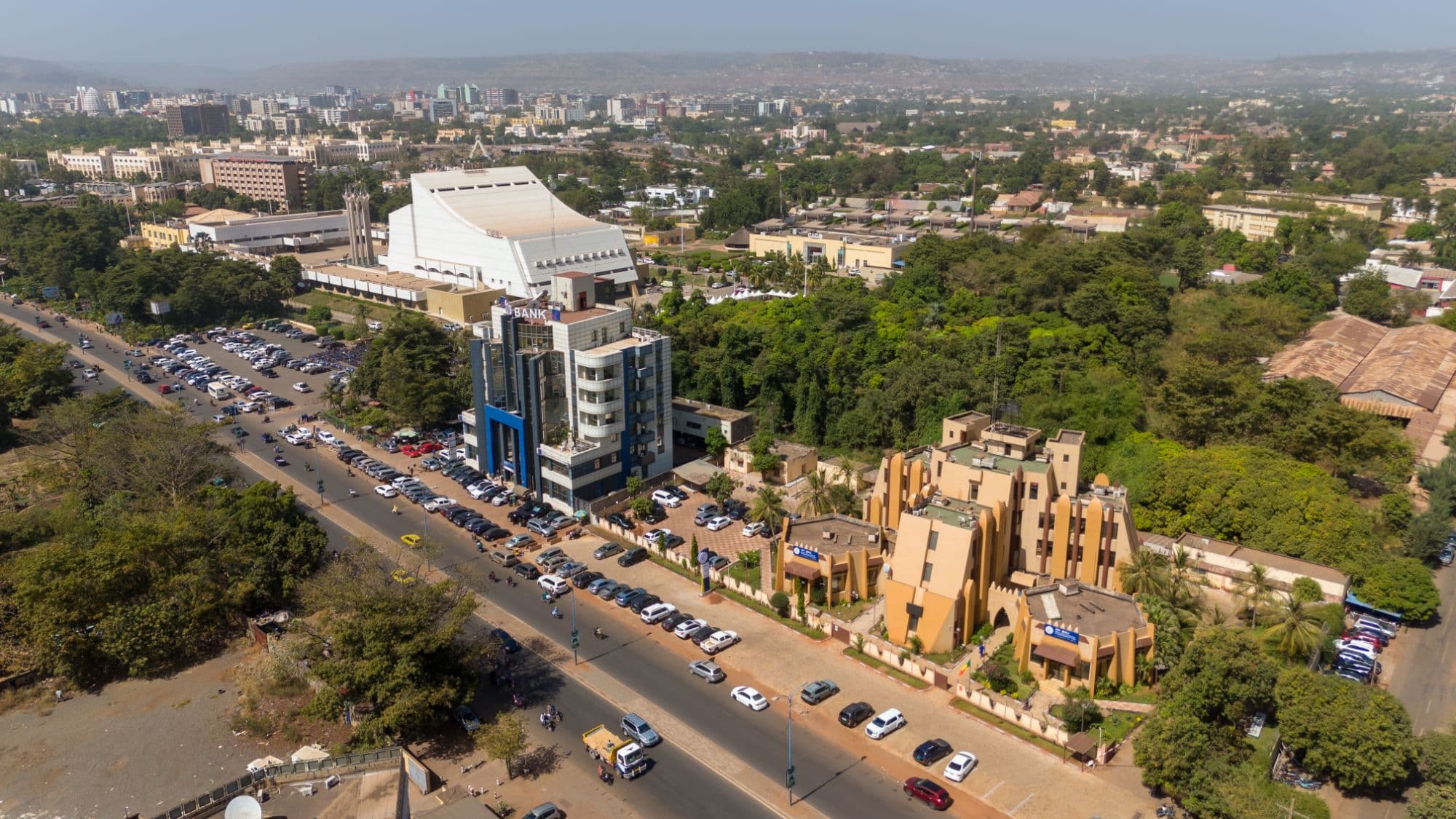 Aerial view of ACI 2000 in Bamako: the International Conference Centre, a large white building with a sloping roof, above a boulevard lined with parked cars, a blue-and-white office block and an ochre Sudanese-style building, set among dense trees, with the city and hills on the horizon.
