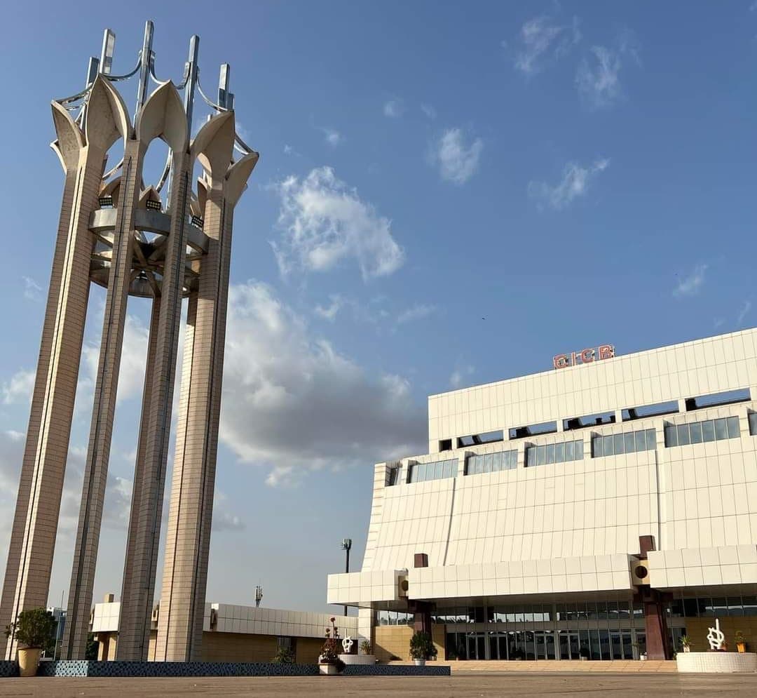 Le Centre international de conférences de Bamako, vu en contre-plongée depuis son parvis : un bâtiment blanc carrelé dont l’étage en porte-à-faux surplombe un socle de pierre sable, l’enseigne orange CICB posée sur la ligne de toit, l’entrée vitrée du rez-de-chaussée derrière des piliers carrés, des plantes en pot et de petites sculptures sur le parvis pavé, et à gauche un haut monument de quatre piliers de béton portant des formes blanches en pétales.