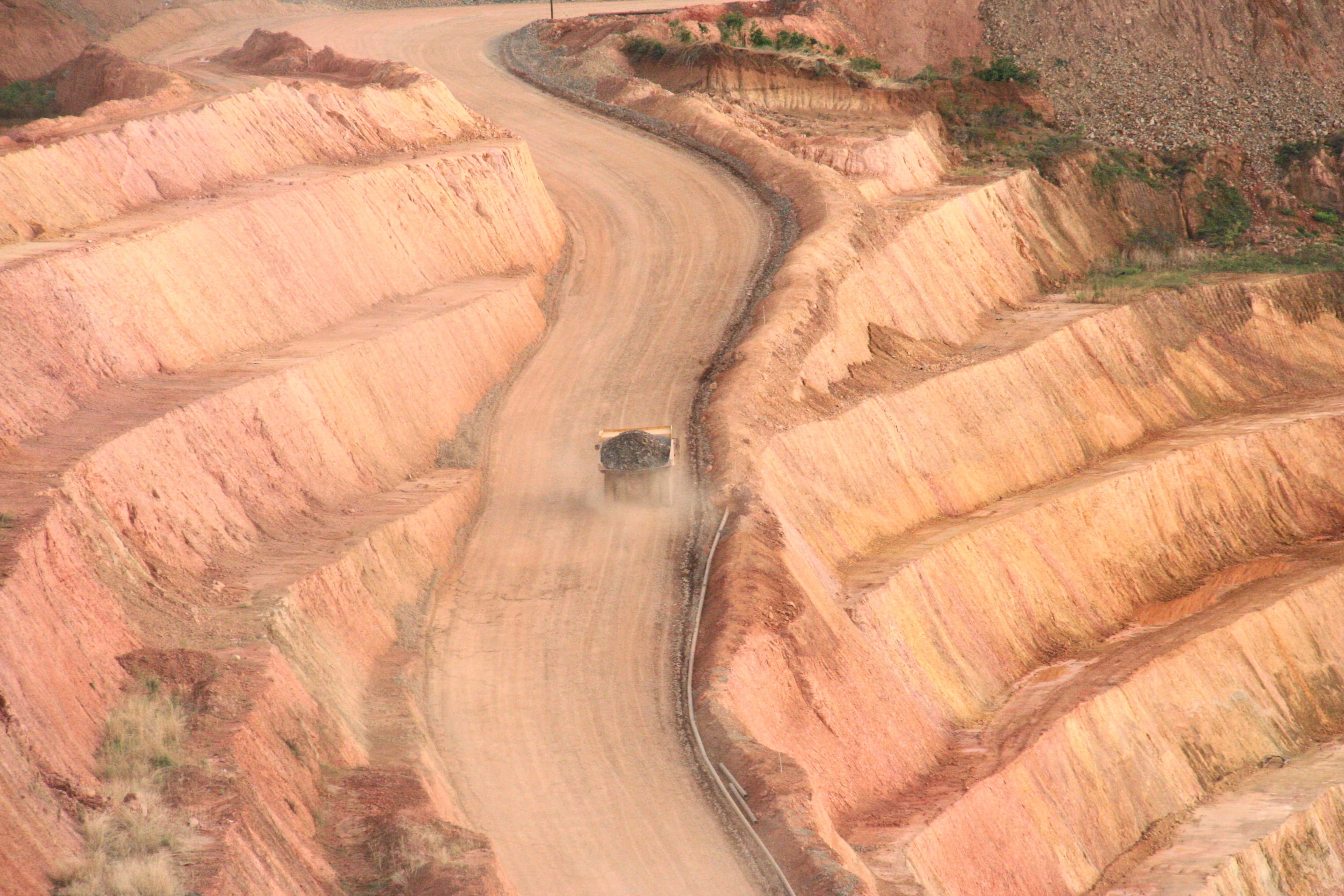 Un camion-benne chargé de minerai gravit une piste damée entre les gradins rose pâle d’une mine à ciel ouvert en Guinée, soulevant un nuage de poussière. — Photo: darrylkeith, "Truck hauling ore from Karta Pit, SMD Crew Gold", via Wikimedia Commons, CC BY-SA 3.0 (https://creativecommons.org/licenses/by-sa/3.0)