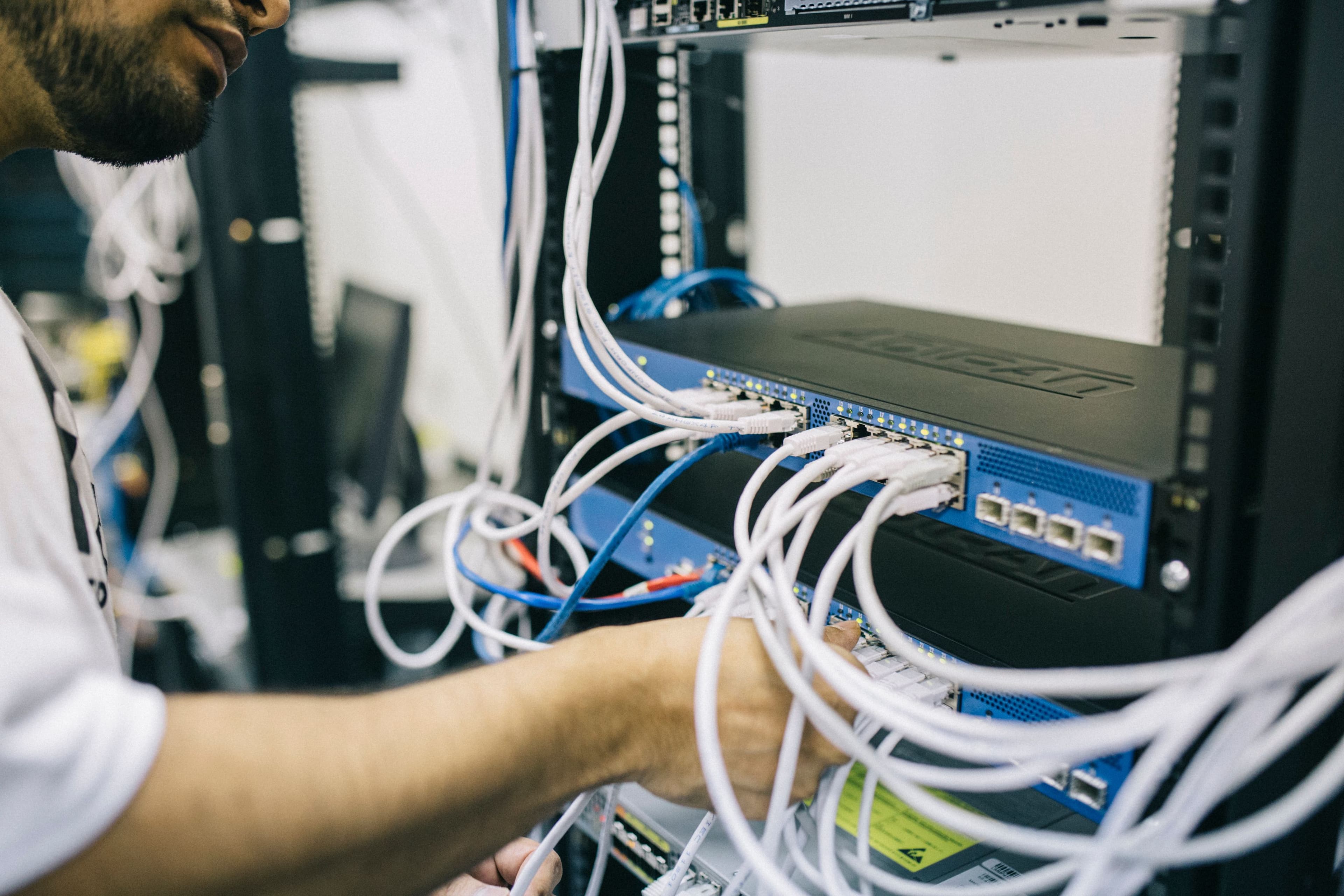 Un technicien habillé de blanc raccorde des câbles Ethernet blancs sur un commutateur réseau bleu monté dans une baie noire, voyants verts allumés sur les ports. — Photo: Field Engineer, "Electronics engineer fixing cables on server", via Pexels, Pexels License (https://www.pexels.com/license/)