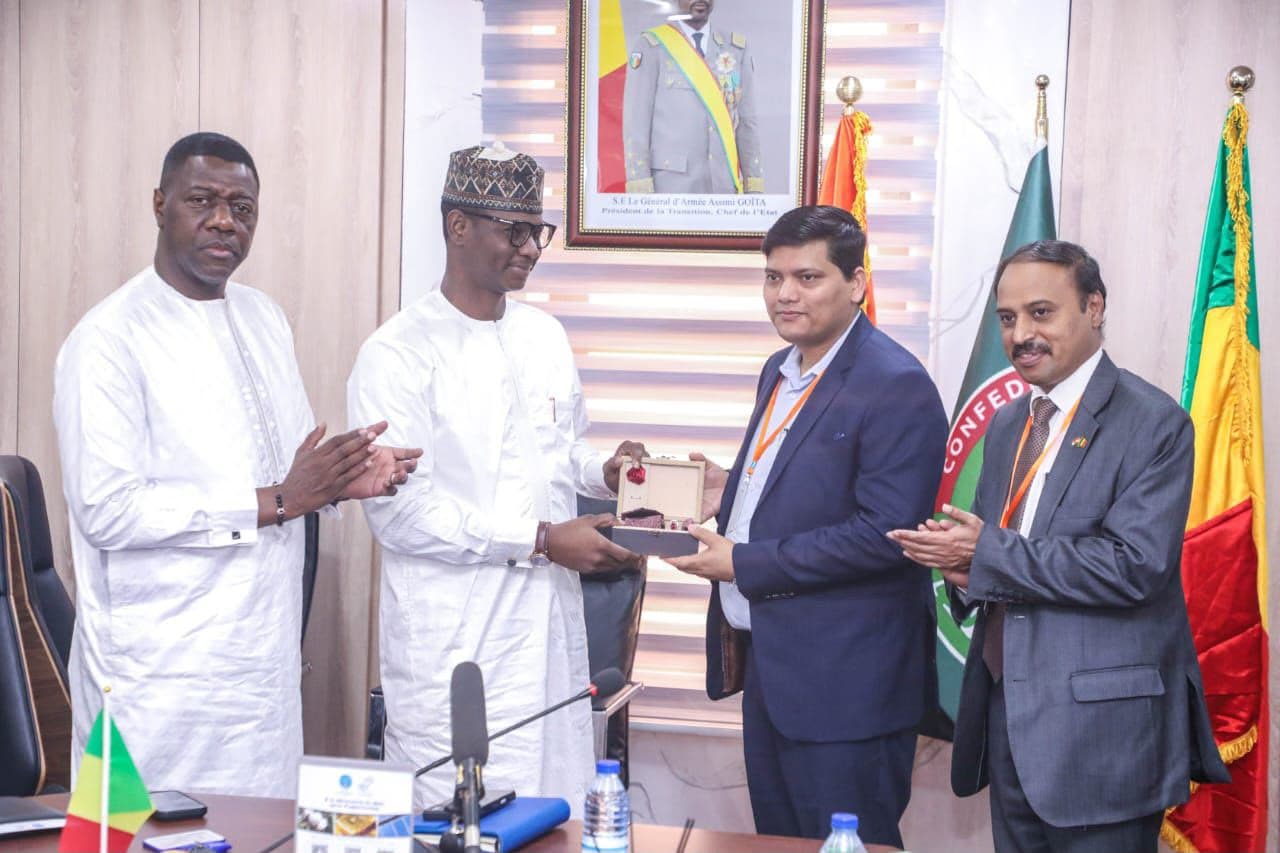 A gift box being presented at an official meeting: two men in white grand boubous and two men in suits wearing orange lanyards, in front of the official portrait of the head of state and the flags of Mali and the Confederation of Sahel States.
