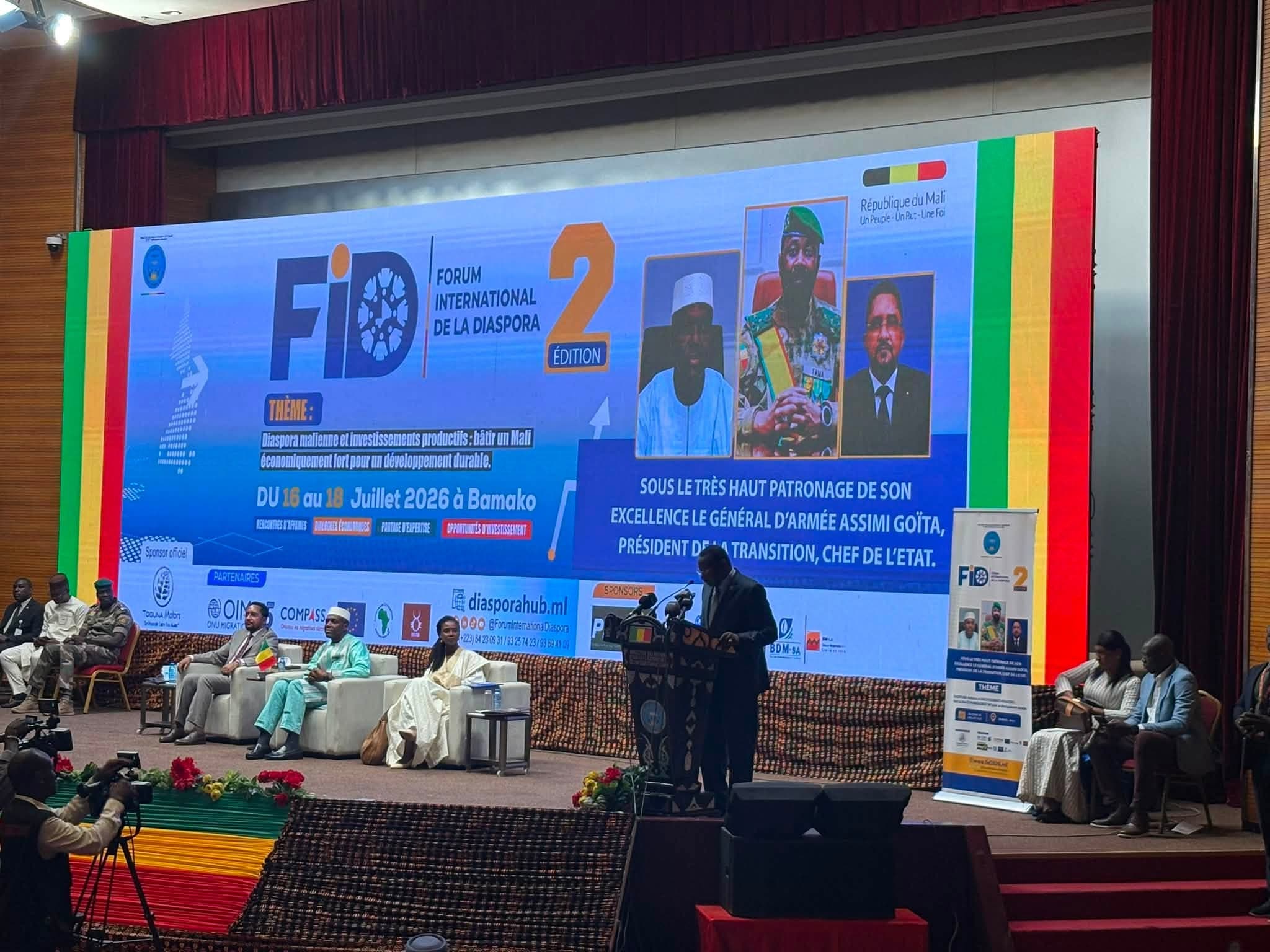 Scène d’un grand auditorium : un orateur au pupitre devant un très large écran LED, trois panélistes assis dans des fauteuils blancs à gauche et des caméras de presse au premier plan. — API-Mali