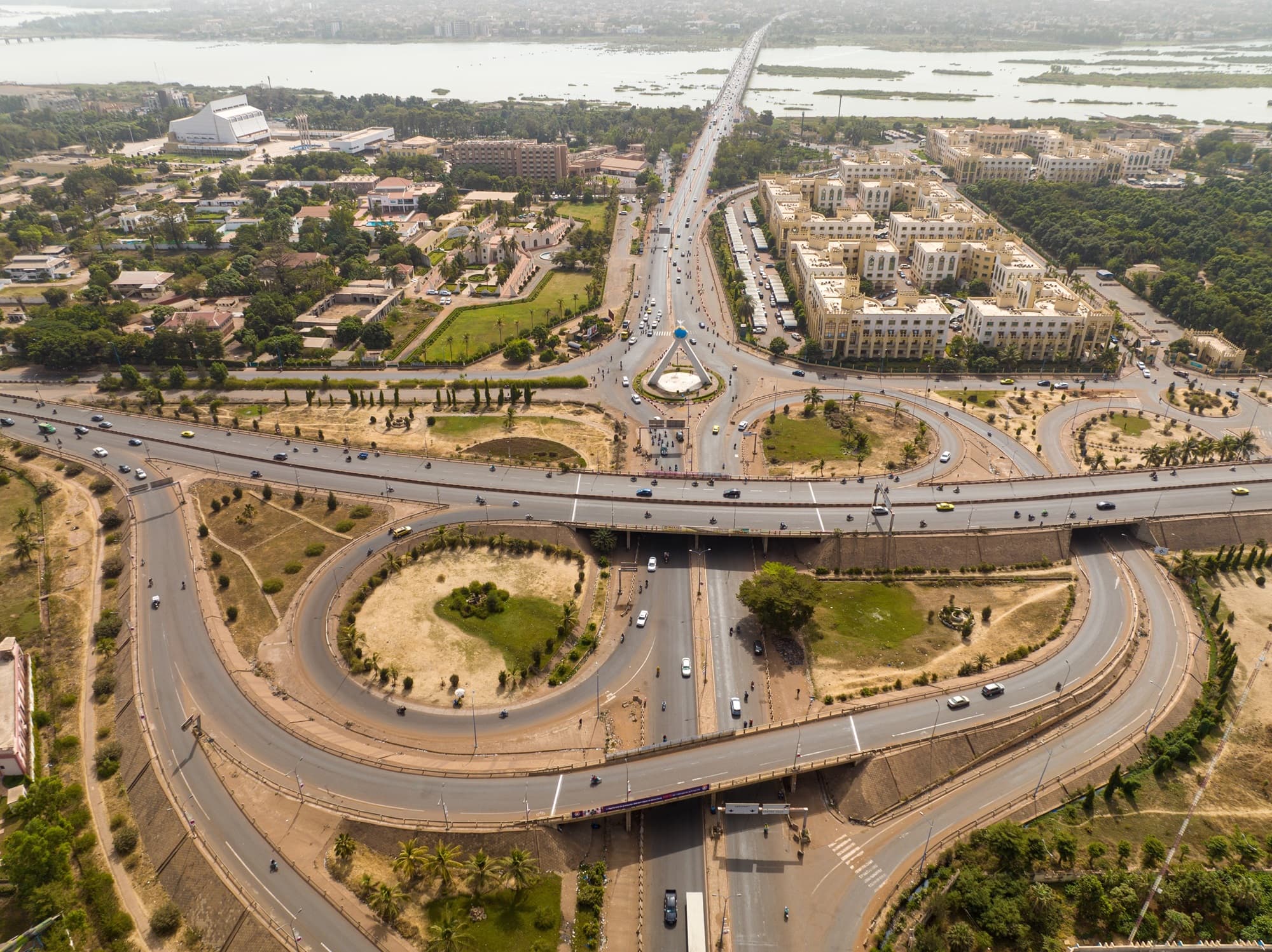 Vue aérienne d’un échangeur à plusieurs niveaux à Bamako, voitures, bus et motos sur les bretelles, un vaste campus administratif beige à proximité et le fleuve Niger avec la ville à l’horizon. — API-Mali