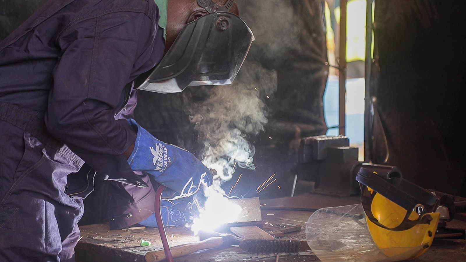 Gros plan sur un soudeur en combinaison bleu foncé, masque et gants de cuir, amorçant un arc sur une pièce d’acier dans un atelier, étincelles et fumée autour de lui. — API-Mali