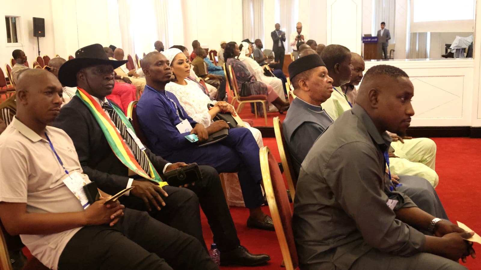 Public assis dans une salle de conférence à moquette rouge, délégués porteurs de badges à cordon, un orateur au pupitre et un photographe au fond de la salle. — API-Mali