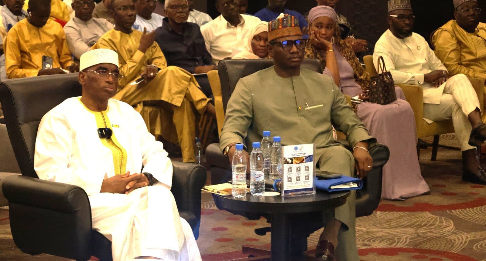 Premier rang d’un public de conférence : deux hommes en boubou assis dans des fauteuils noirs derrière une table basse, des rangées de délégués derrière eux. — API-Mali