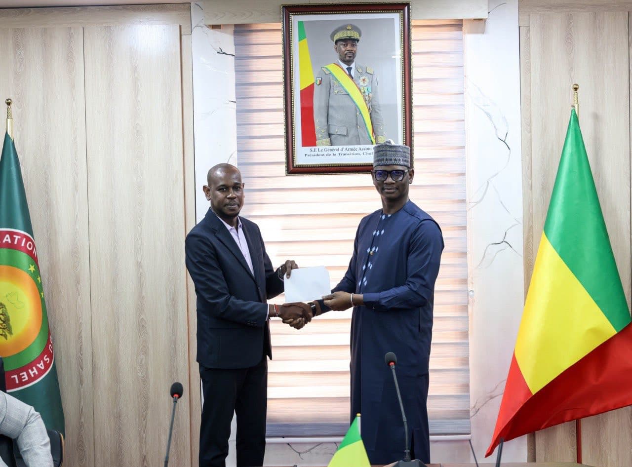 Deux hommes se serrent la main en tenant ensemble une enveloppe blanche dans une salle de réunion officielle lambrissée, drapeaux et portrait officiel encadré au mur. — API-Mali